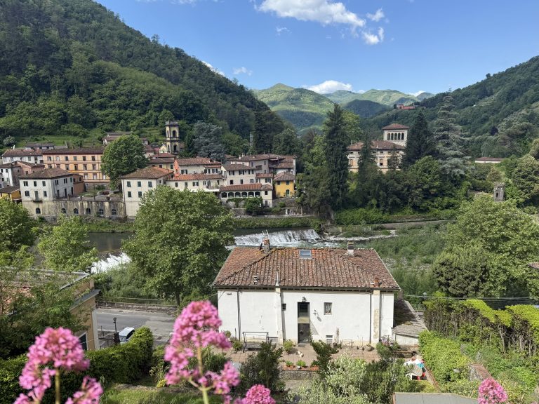 View of Bagni di Lucca with terracotta rooftops, a river, and a small waterfall surrounded by green hills