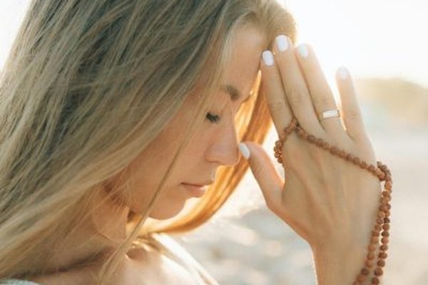Woman with long blonde hair and eyes closed, holding a string of mala beads with an orange tassel to her forehead in soft sunlight