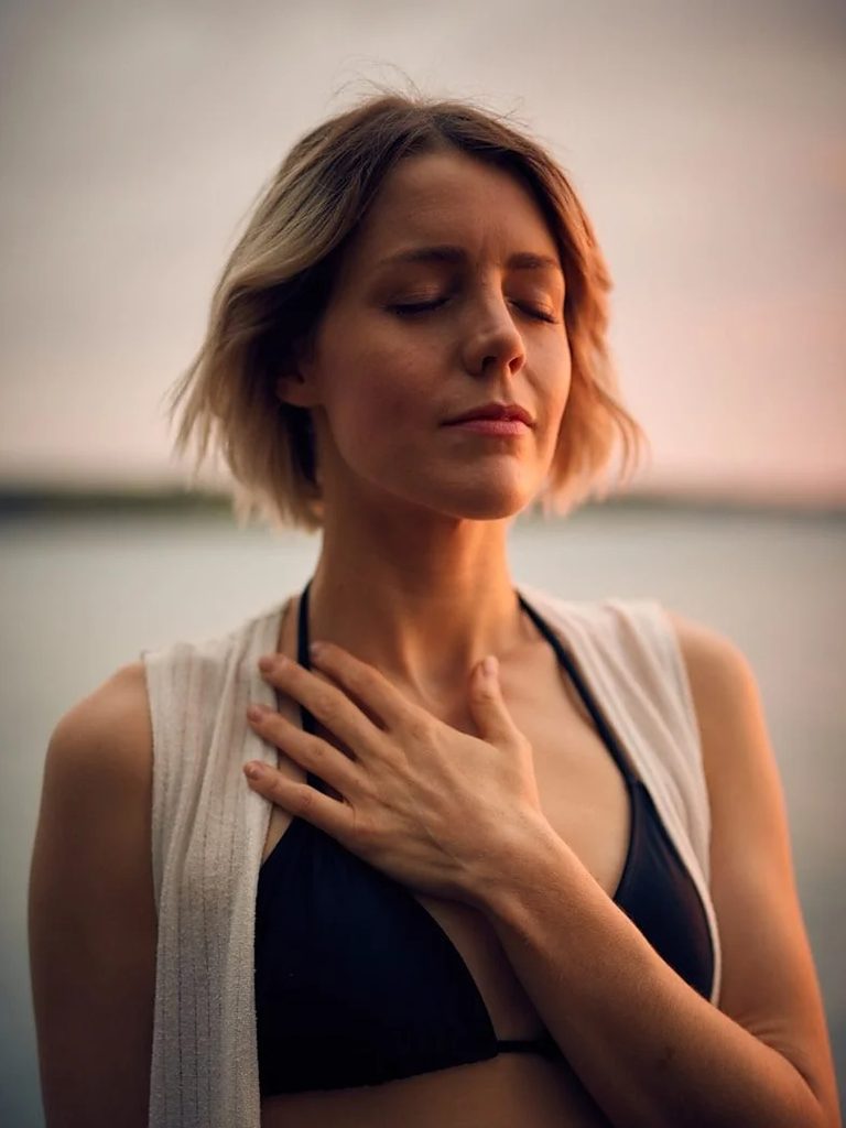 Woman with eyes closed and hand on her chest, breathing calmly in soft golden evening light by the water