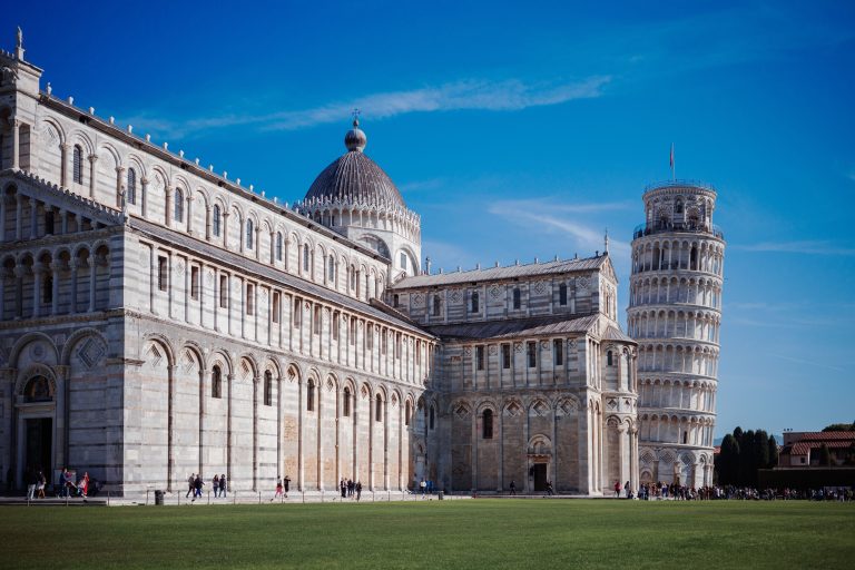 White marble Pisa Cathedral with its grey dome and the Leaning Tower of Pisa beside it, green lawn in front and a clear blue sky above