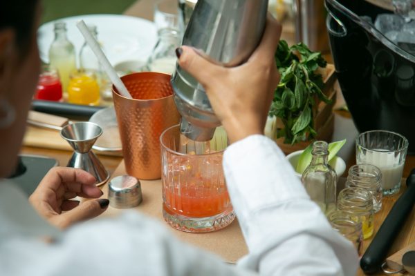 Bartender in a white shirt pouring from a cocktail shaker into a glass of red drink, with a copper mug, jigger, fresh mint, and small bottles of syrup on the table
