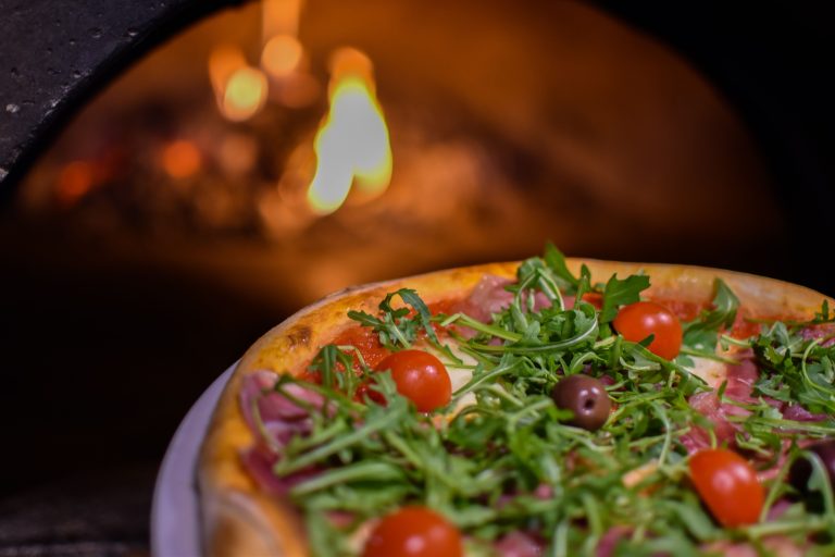 Pizza topped with prosciutto, rocket, cherry tomatoes, and olives in front of a glowing wood-fired oven