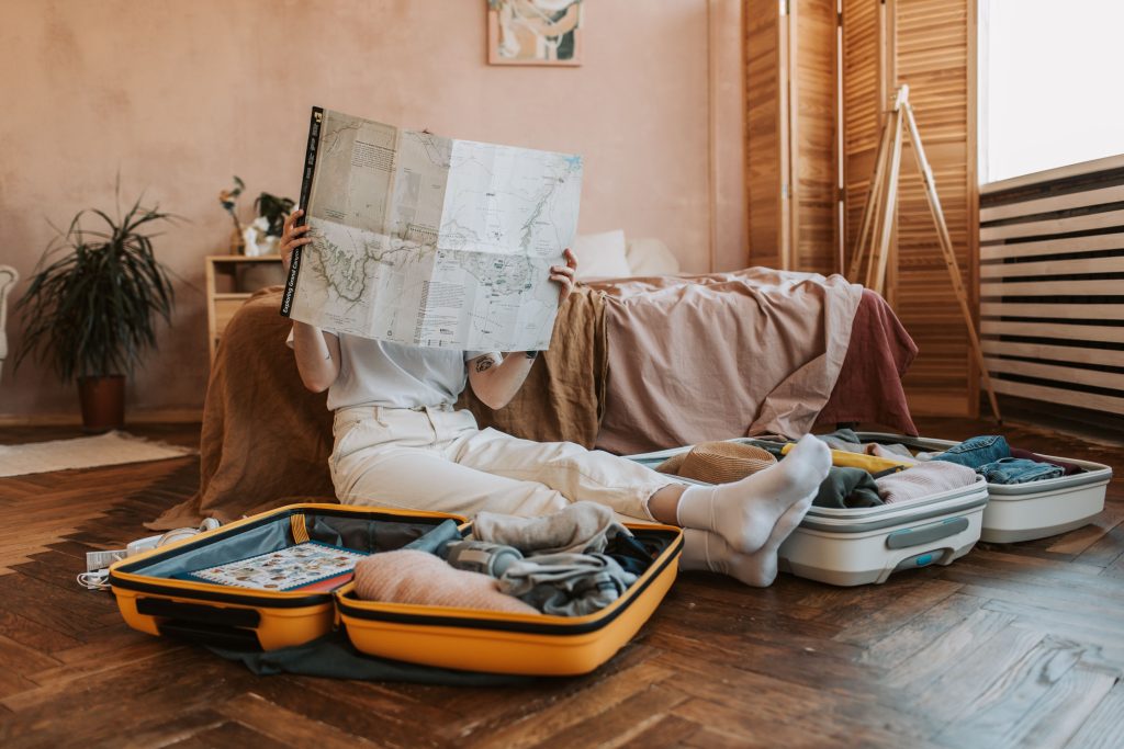 Woman in white socks sitting on a wooden floor between two open suitcases, holding up a large map that hides her face